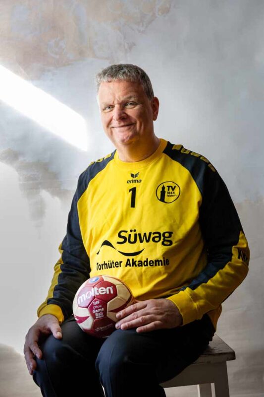 Dr. Wagner sitzt im Trikot eines Handballtorhüters mit einem Ball und lächelt in die Kamera.